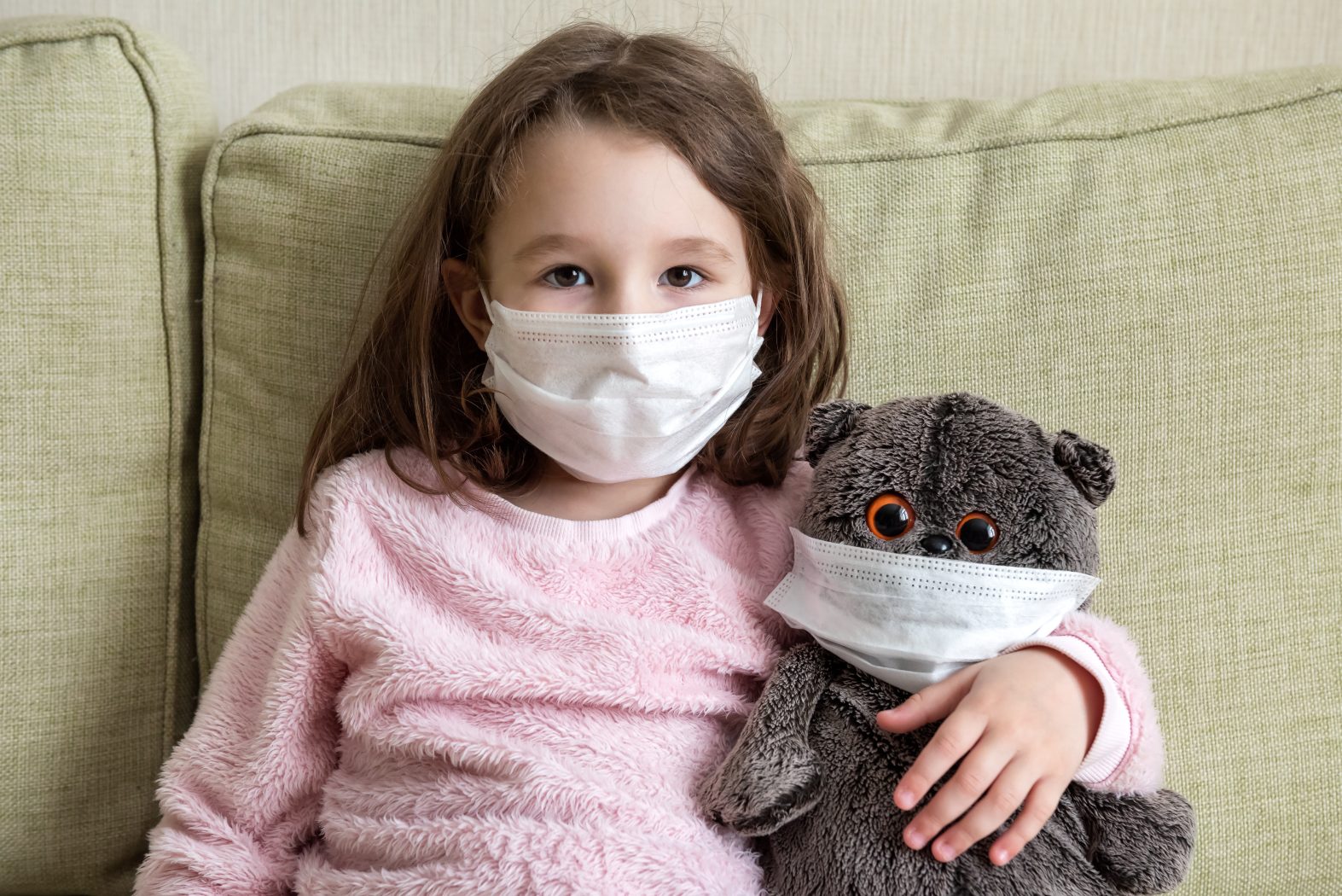 tay at home in quarantine, funny kid with toy on couch during COVID-19 coronavirus pandemic. Little girl in medical mask for protection to corona virus indoors. Safe live and coronavirus concept.