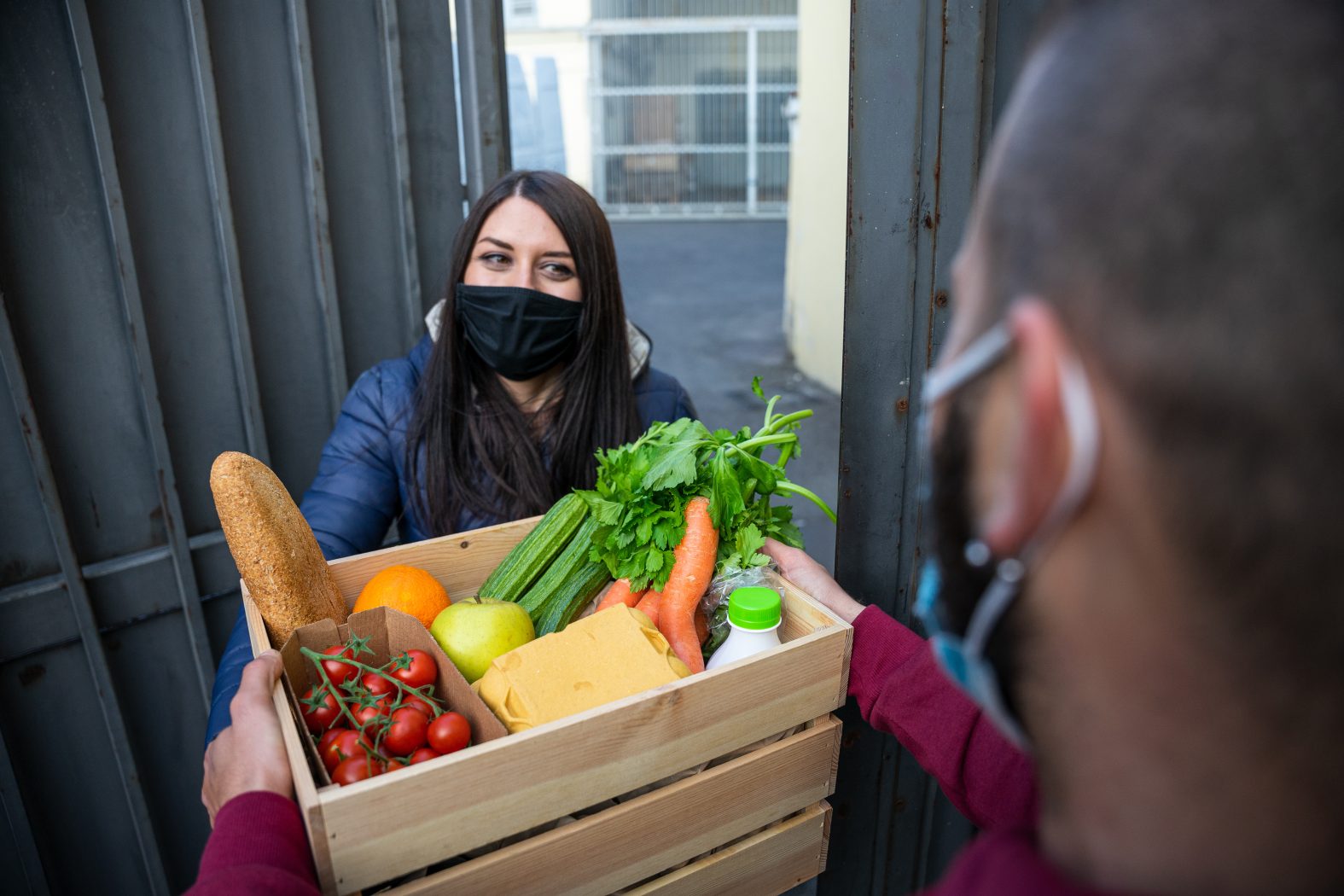 Young man courier delivers online shopping basket with fresh fruit vegetables to woman customer's home during Coronavirus Covid-19 pandemic quarantine wearing protective face mask - Concept delivery