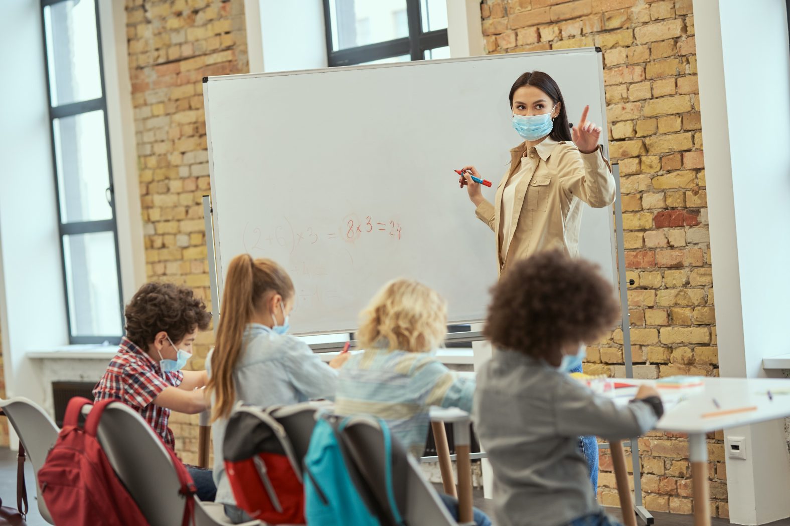Female teacher wearing protective mask during coronavirus pandemic writing mathematic task on board for group of elementary school kids. Education, math and covid19 concept